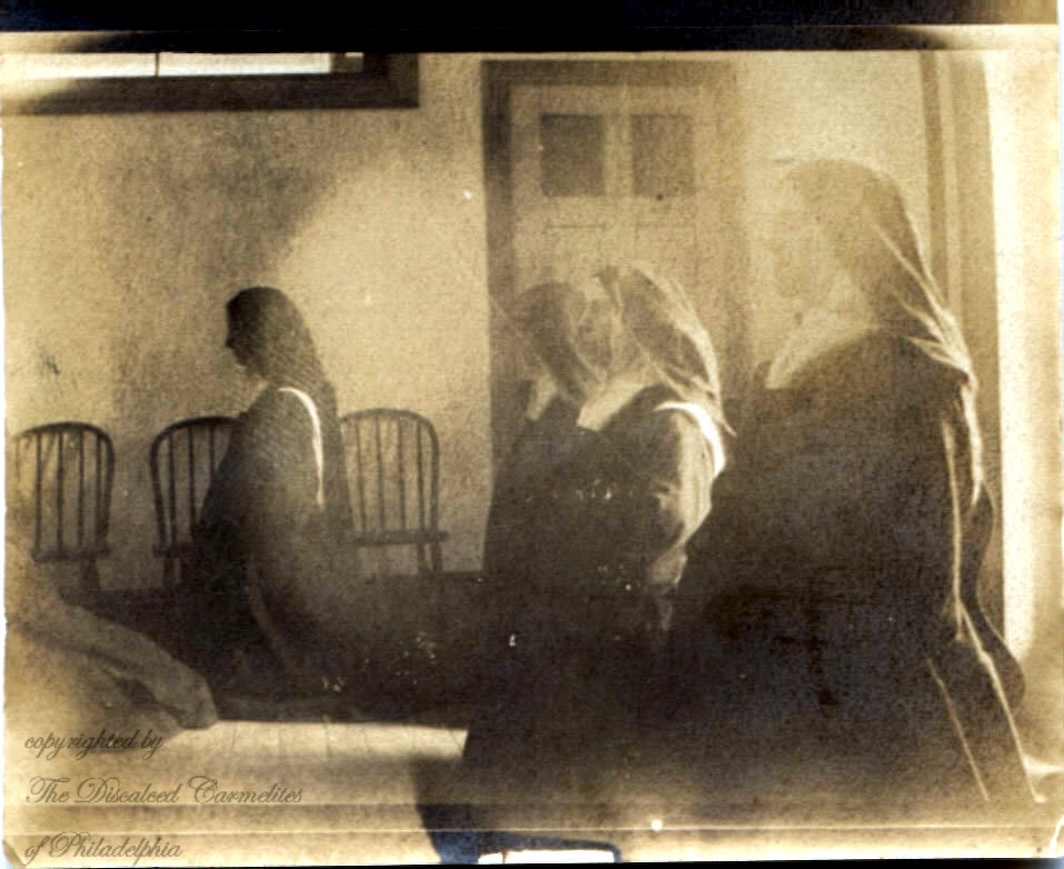 sepia photo of four Carmelite nuns in habit kneeling in a chapel with plain white walls and a wood floor, shot from the side