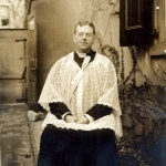 Father Moore in black cassock and white alb, seated, photographed outside, perhaps outside the Carmel