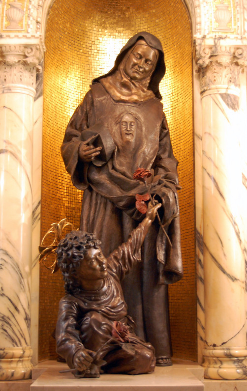 bronze statue stands in niche of chapel against gold background.  St. Therese has an image of the Holy Face emblazoned on her breast.  In her right hand she holds the doctoral biretta.  A child on the ground reaches up, offering her a rose.