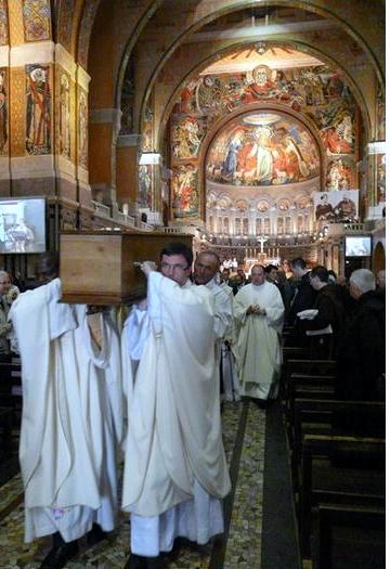 color photo of priests in white vestments carrying Bishop Gaucher's coffin out of the Basilica of St. Therese in Lisieux