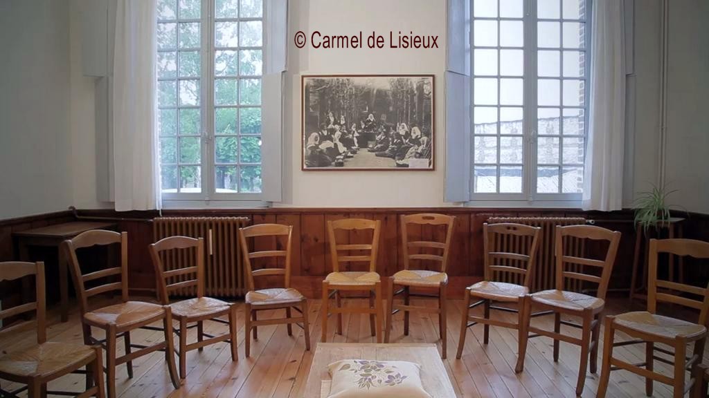 empty recreation room, windows looking out into garden of Lisieux Carmel, straight chairs in a semicircle, photo of Therese with community at recreation in the chestnut walk hangs on white wall between windows