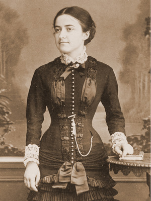 formal black and white photo of Pauline Martin as a young laywoman, wearing a dark suit with elaborate buttons and ruching, white lace collar and cuffs, resting her hand on a stack of books on a table